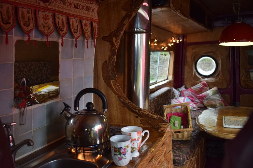 Compact, rustic caravan interior with wooden accents. Foreground: kitchenette with a stainless steel sink, kettle on a small stove, two floral mugs on the counter. Background: cozy dining nook with cushions and fairy lights around a small round table. Round window offers natural light.