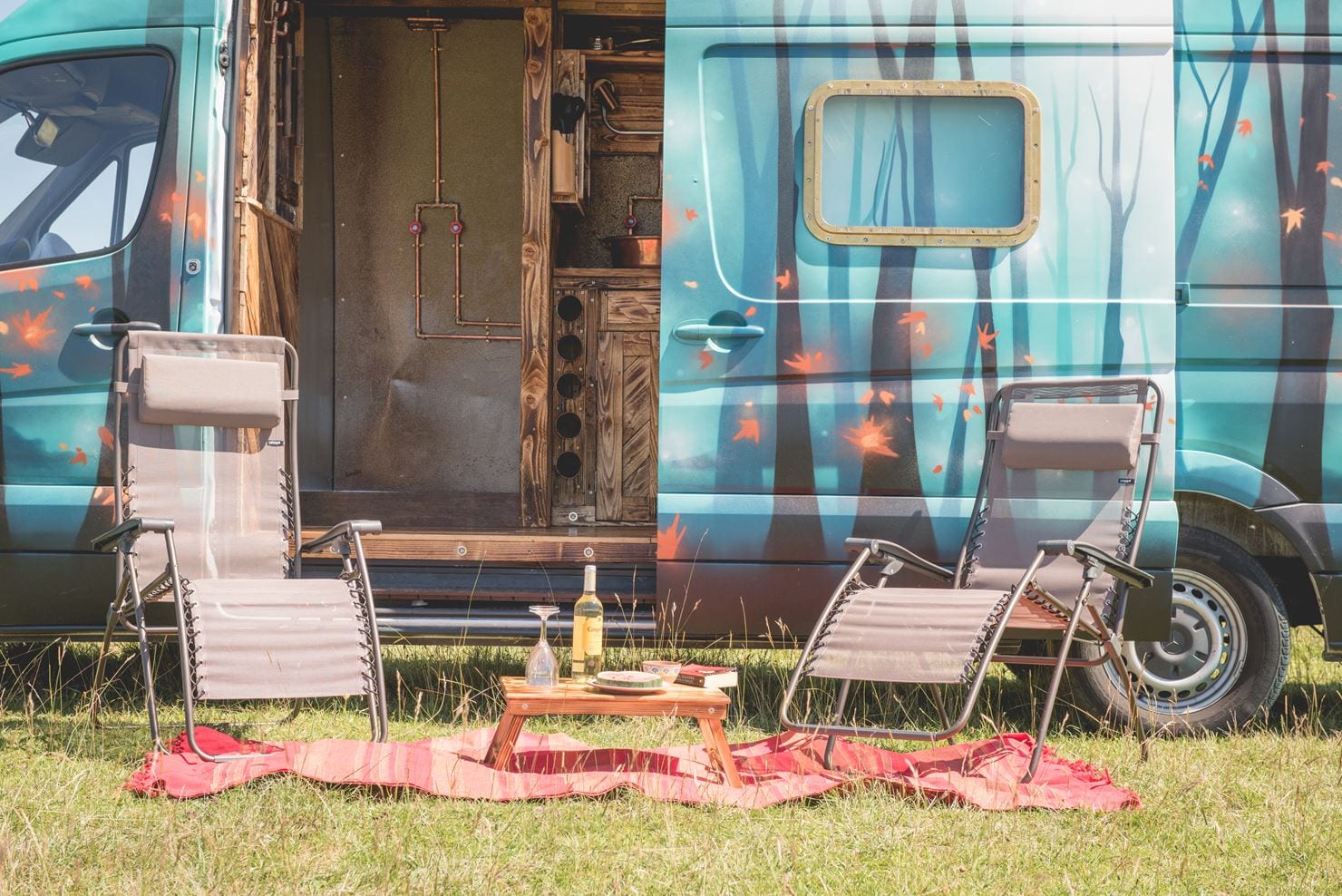 The outside of a campervan with two picnic chairs, wine and a picnic blanket
