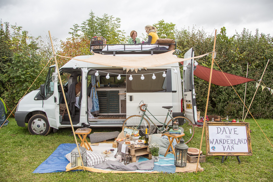 a converted ford transit campervan wih two people relaxing on the roof and rugs and chairs in front