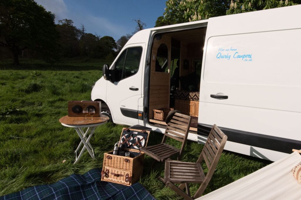 Outside of a campervan  with picnic bx, blanket and two chairs 