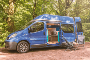 A blue campervan with two camp chairs parked in a wooded area