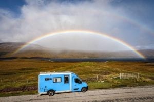 A blue campervan drives along a road next to a loch in Scotland with a rainbow in the background