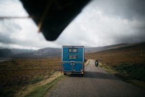 The view of the back of a blue campervan from a car with a surfboard on the roof  on an empty road in the highlands of Scotland