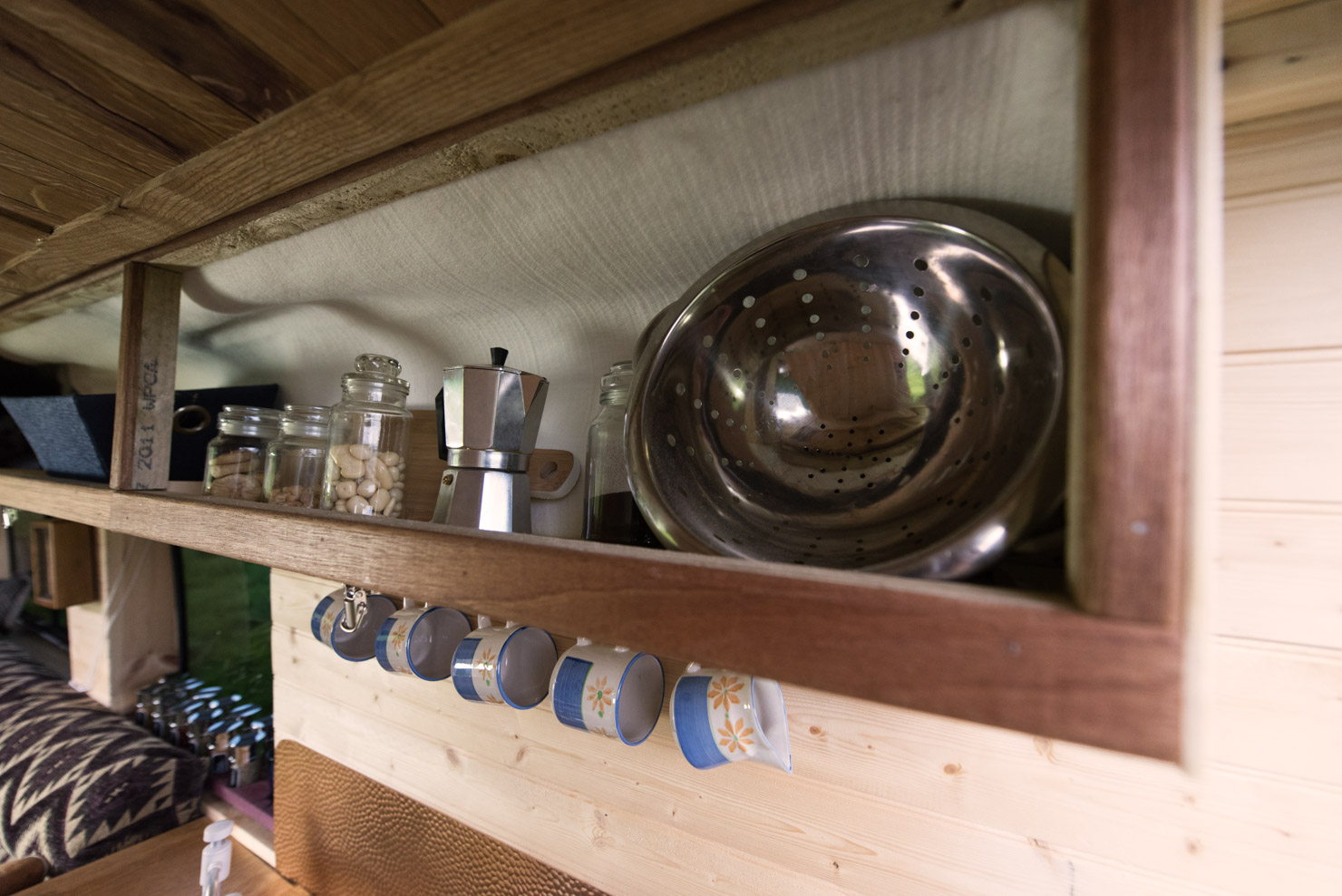 A wooden shelf inside a compact kitchen area holds several items, including a metal colander and glass jars with food. Below the shelf, mugs hang from hooks. The background features light-colored wooden paneling and a patterned cushion on the left side.