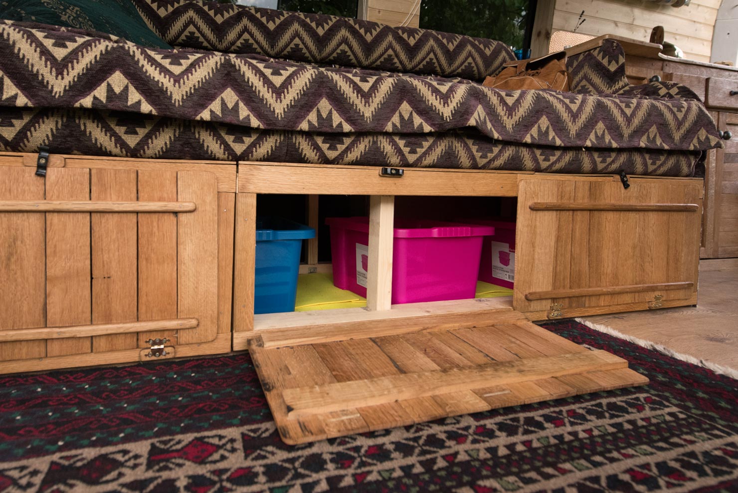 A wooden storage compartment under a cushioned seating area is open, revealing a blue plastic bin and a pink plastic bin inside. The seating has a patterned fabric with earthy tones. The wooden compartment has two hinged doors and a drop-down panel. A patterned rug is visible in the foreground.