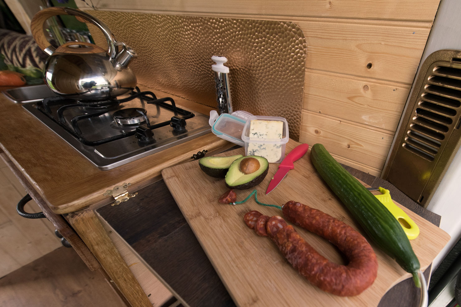 A kitchen counter with a wooden wall in the background. On the counter are an avocado, sliced sausage, a cucumber, a knife with a red handle, and a small container of blue cheese. A stainless steel teapot sits on a gas stove burner to the left, and a yellow peeler is next to the cucumber.