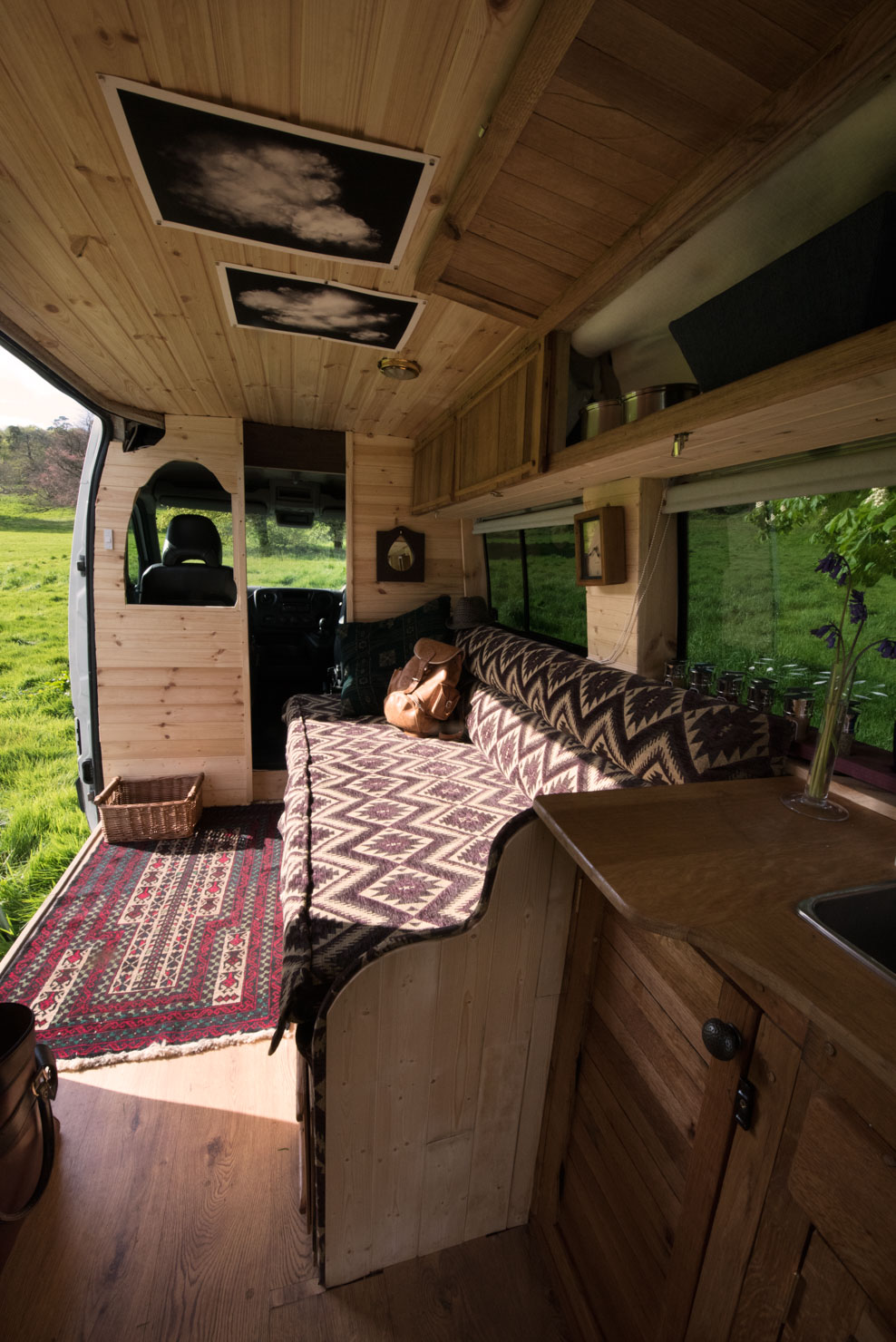 Interior of a cozy campervan with wooden paneling. The space features a patterned sofa, a woven rug, and a small kitchen area with a sink. A teddy bear sits on the sofa. Sunlight filters in through the open side door, revealing a green field outside. Photos and storage shelves adorn the walls.