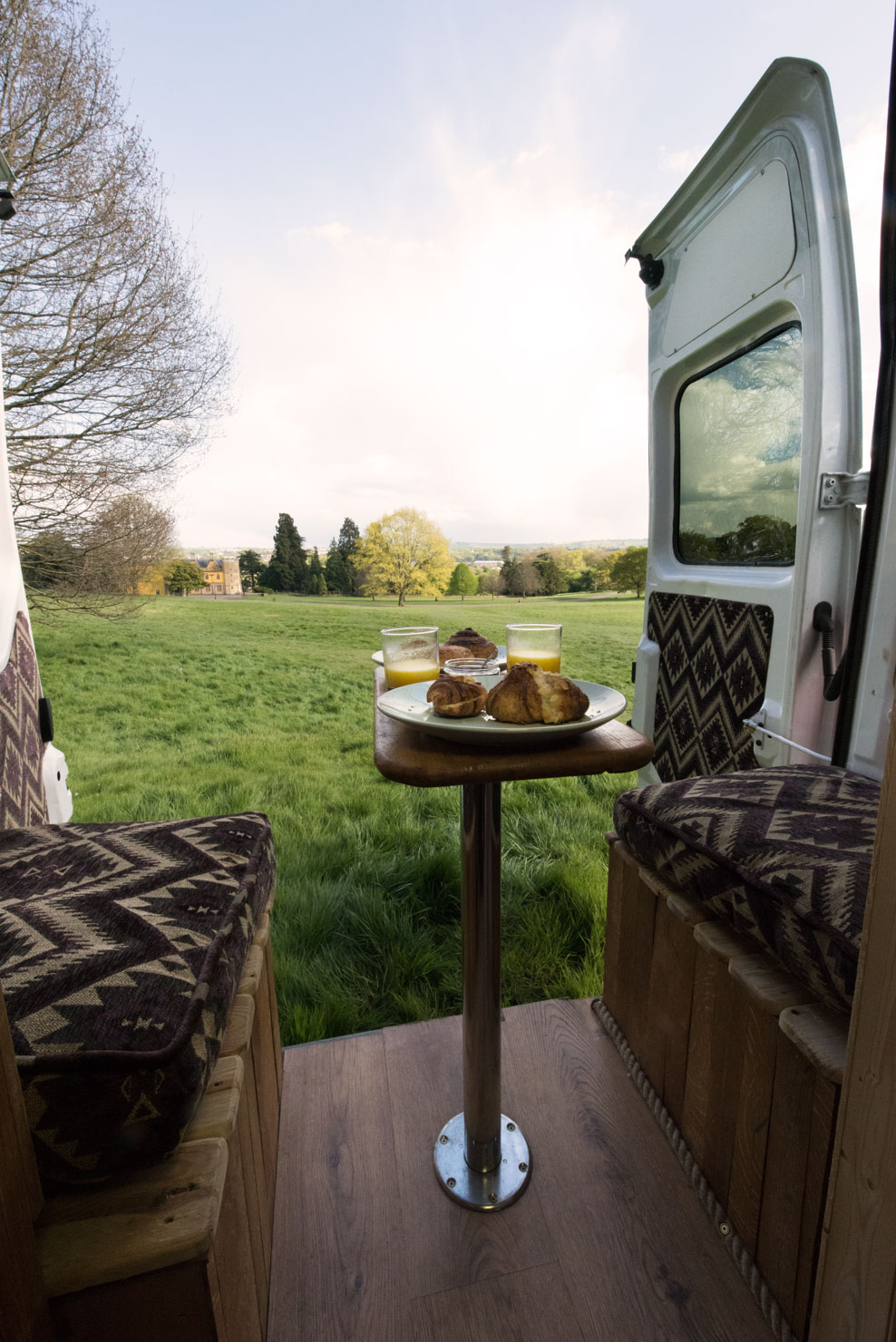 Interior view of a campervan's open back door overlooking a grassy field. Inside the van, a small wooden table is set with plates of pastries and glasses of juice. The van has cushioned seating with patterned upholstery. The background features trees and a partly cloudy sky.