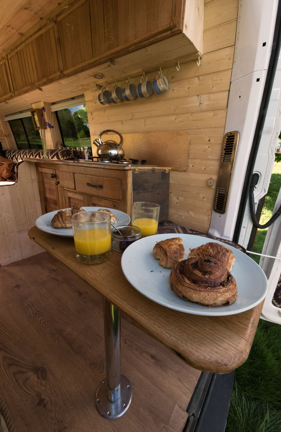 A campervan interior with wooden walls and flooring is shown. A small fold-out table holds two white plates, each with pastries, and two glasses of orange juice. In the background, a countertop with a kettle, hanging cups, and a window with a scenic view is visible.