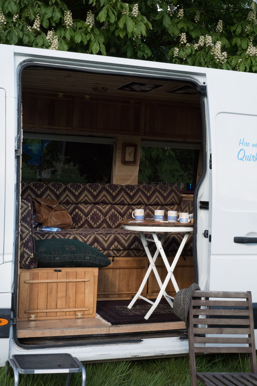 An open white camper van reveals a cozy, rustic interior with wood paneling. Inside, there's a small foldable white table with teacups and plates, a patterned couch with a brown bag on it, and a cushion on a storage box. A folding chair is placed outside near the grassy ground. Trees are visible in the background.