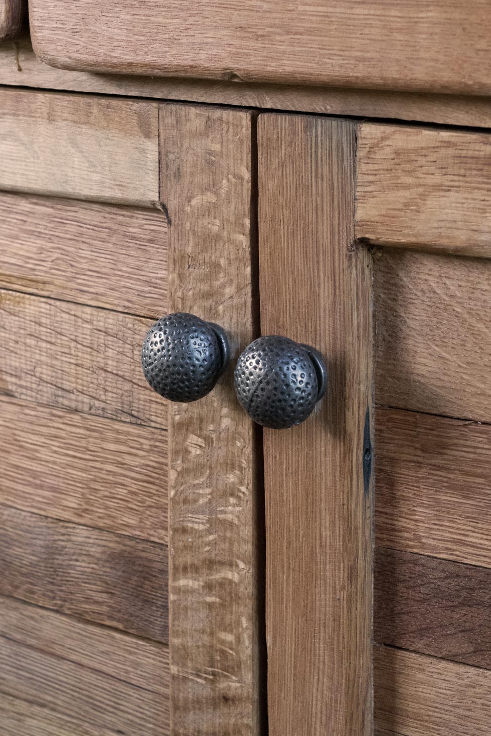 Close-up image of a wooden cabinet with two textured round metal knobs. The wood has a slightly rough, rustic finish with a natural brown hue and visible grain patterns. The knobs are spherical, dark, and feature a dimpled surface, adding an antique or vintage look to the cabinet.