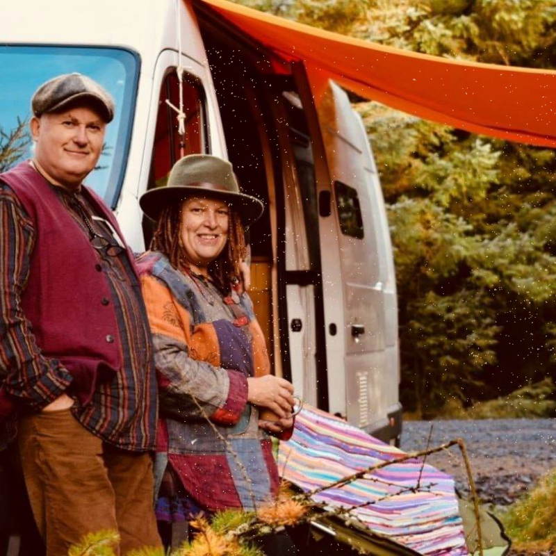 A man and woman stand beside a van with its door open. Both are wearing hats and colorful, casual clothing. A striped cloth is draped near the van and an orange sheet is tied overhead, suggesting an outdoor setup. The background is filled with green foliage, indicating a forest setting.