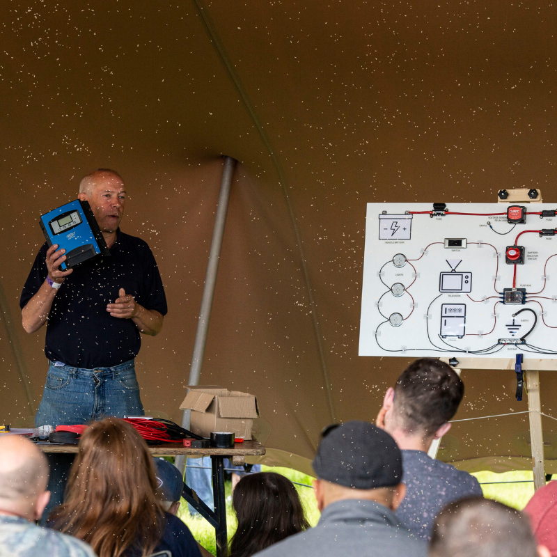A man stands next to a whiteboard with electrical diagrams, holding a blue device. He is giving a presentation under a tent to a seated audience. The whiteboard shows a schematic with symbols, arrows, and components connected by lines. Various equipment is placed on the table in front of him.