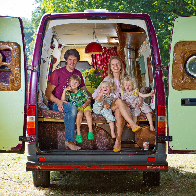 A family of five sits in the back of an open van, surrounded by vibrant greenery. The parents, with three young children, all smile warmly. The van is cozy and colorful, with floral cushions and various decorations. An open door on either side reveals the lush forest background.