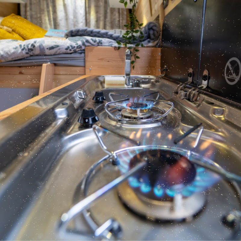A close-up of two gas burners with blue flames on a stainless steel stovetop inside a camper van. In the background, a cozy bed with yellow bedding, a book, and patterned curtains can be seen. A small potted plant hangs near the sink, and the interior shows wood-panel accents.