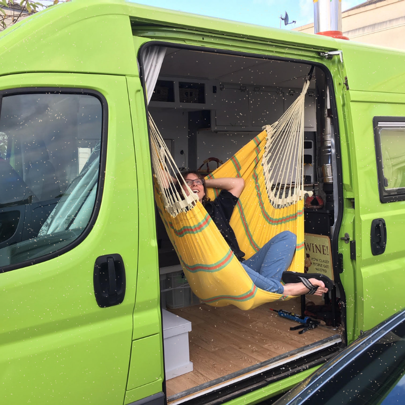 A person lounges in a yellow hammock hung inside a bright green van with its side door open. The van is parked, revealing a wooden interior and various belongings. The individual is relaxed, lying back with one arm behind their head and the other holding a device, possibly a phone.