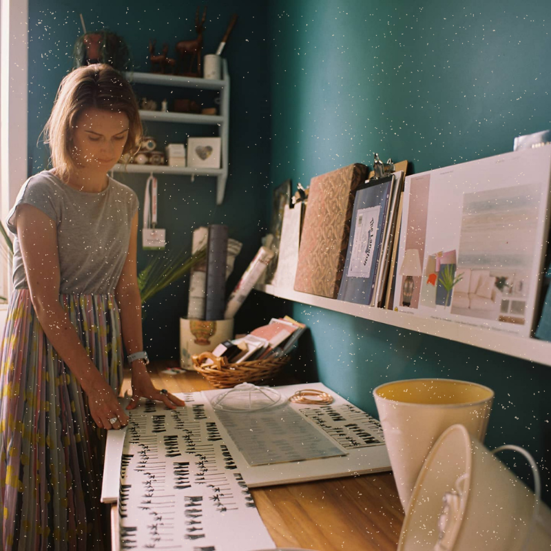A woman in a gray T-shirt and colorful skirt stands at a wooden desk, examining a large sheet with abstract black designs. The room is painted teal and decorated with shelves holding various items, including books, fabric rolls, and art supplies. Sunlight streams in from the left, casting a warm glow.