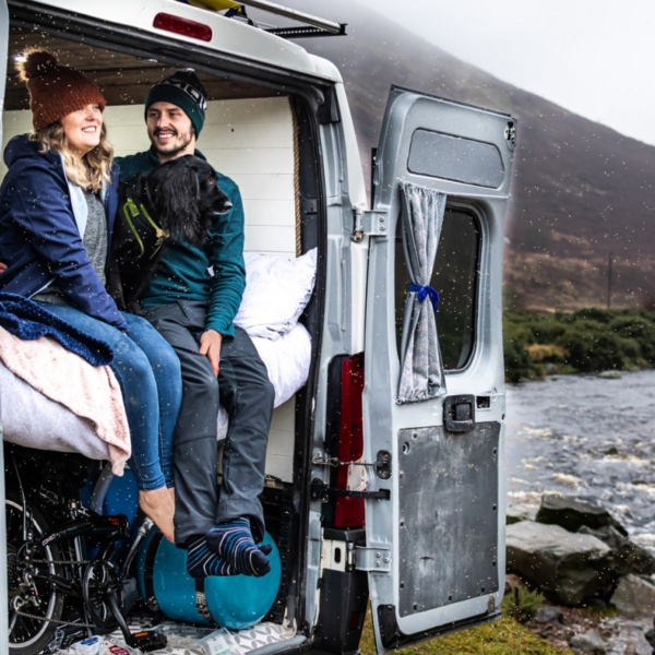 A couple sits at the back of a van converted into a cozy living space near a river. The woman, in a brown beanie and blue jacket, and the man, in a black hat and green jacket, smile while their black dog stands between them. The van door is open, revealing mountains and a river under a cloudy sky.