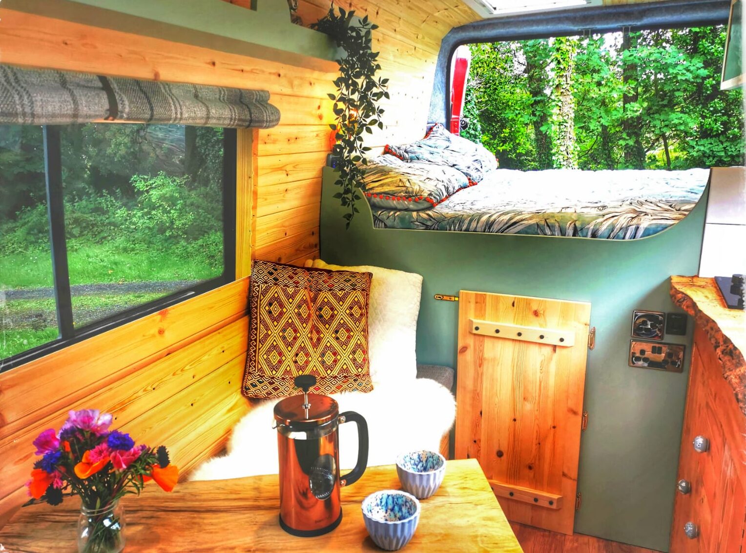 A cozy camper van interior features a small wooden dining table with a French press and two mugs, and a cushioned seating area with patterned pillows and a throw blanket. A raised bed is in the background near a window with green trees outside. A vase of colorful flowers sits on the table.