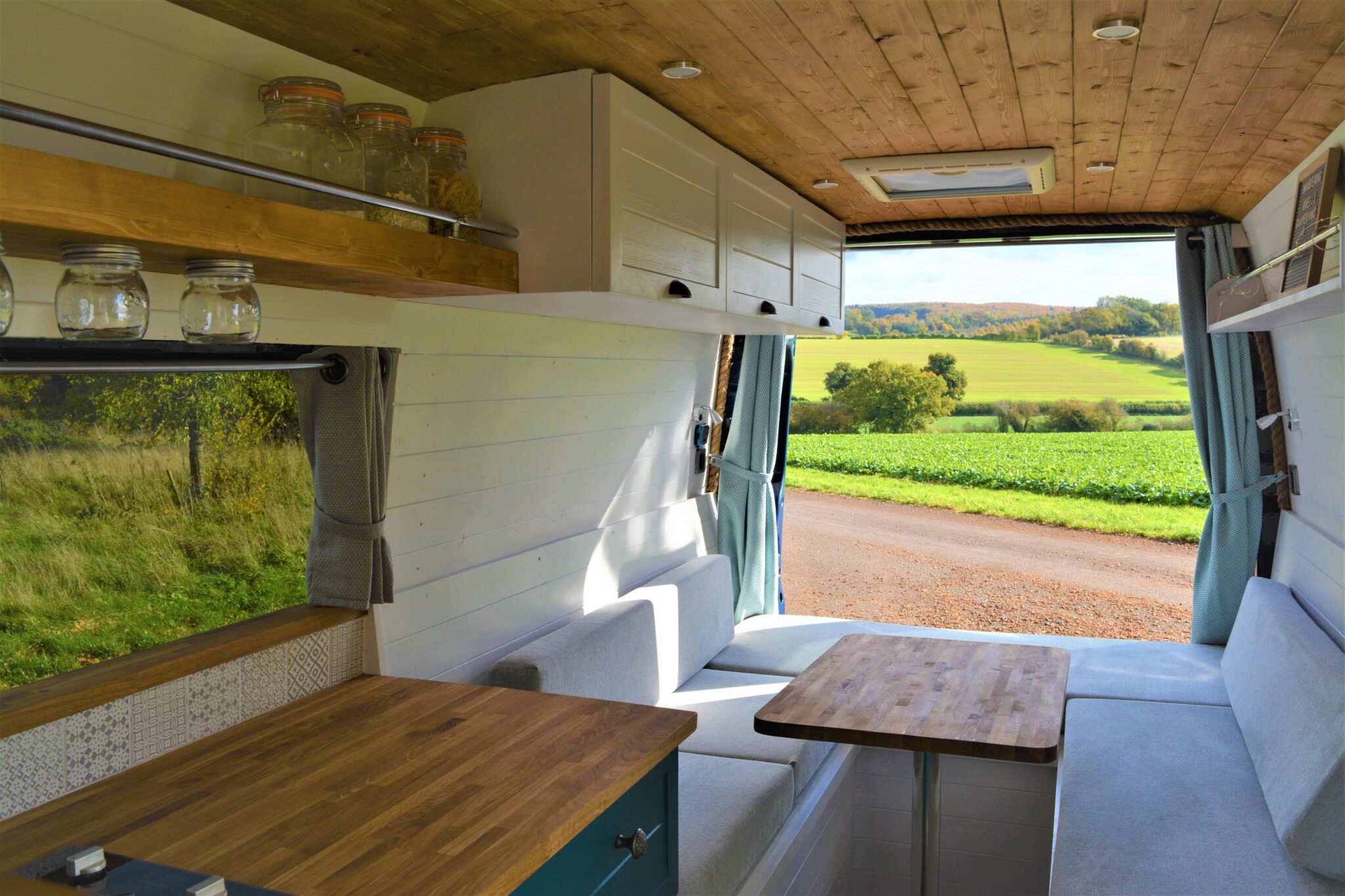 Interior of a camper van with wooden countertops and white cabinetry. A seating area with a wooden table faces large windows, offering a view of a green, rolling countryside. Glass jars are stored on a wooden shelf. Blue curtains frame the open rear doors and sunlight fills the space.