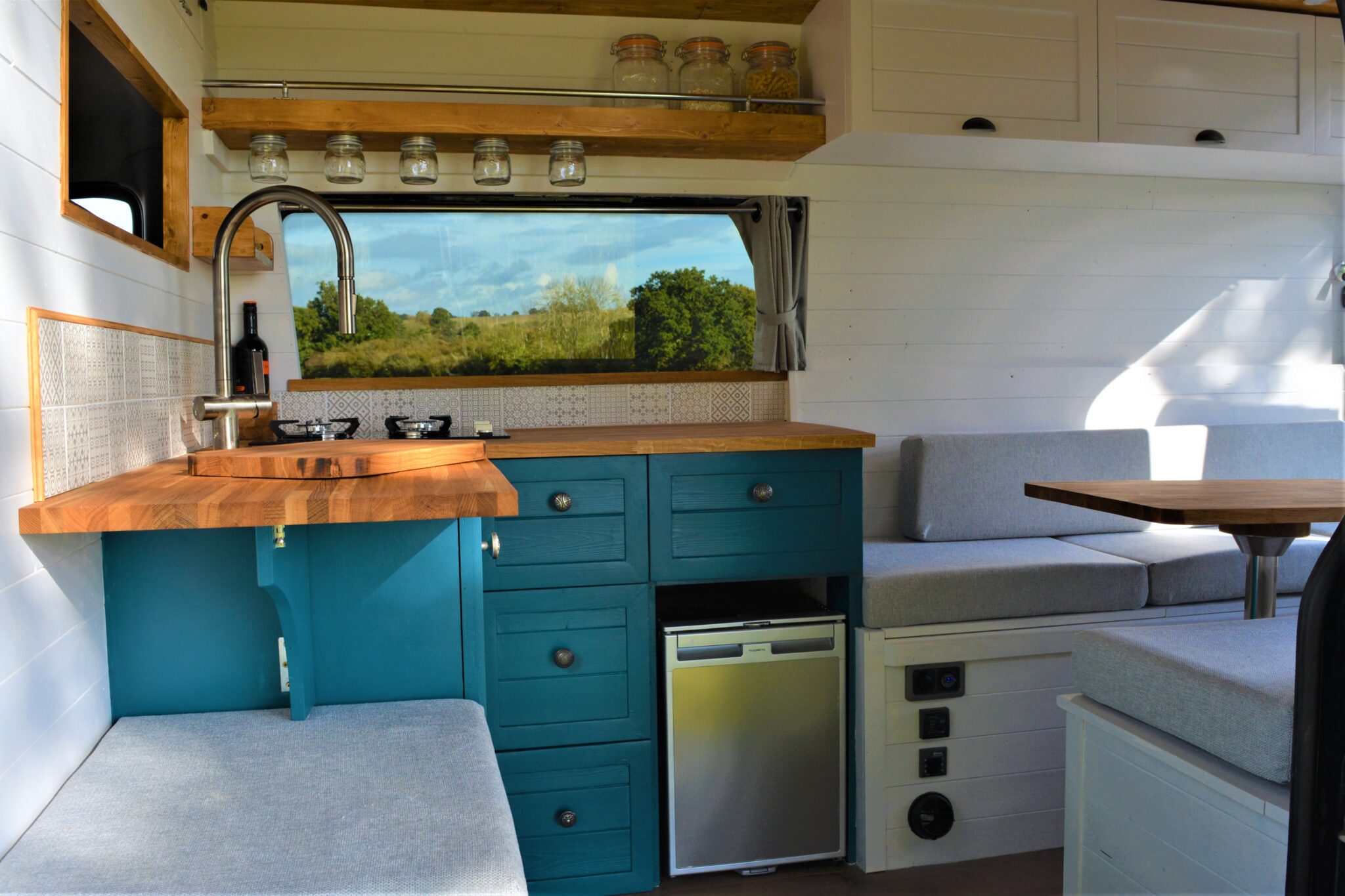 Interior of a camper van featuring a compact kitchen with blue cabinets, a wooden countertop, and a stainless steel sink. There are shelves with mason jars above the window. A mini-fridge is below the counter. Adjacent is a cozy sitting area with gray cushions and a small wooden table.