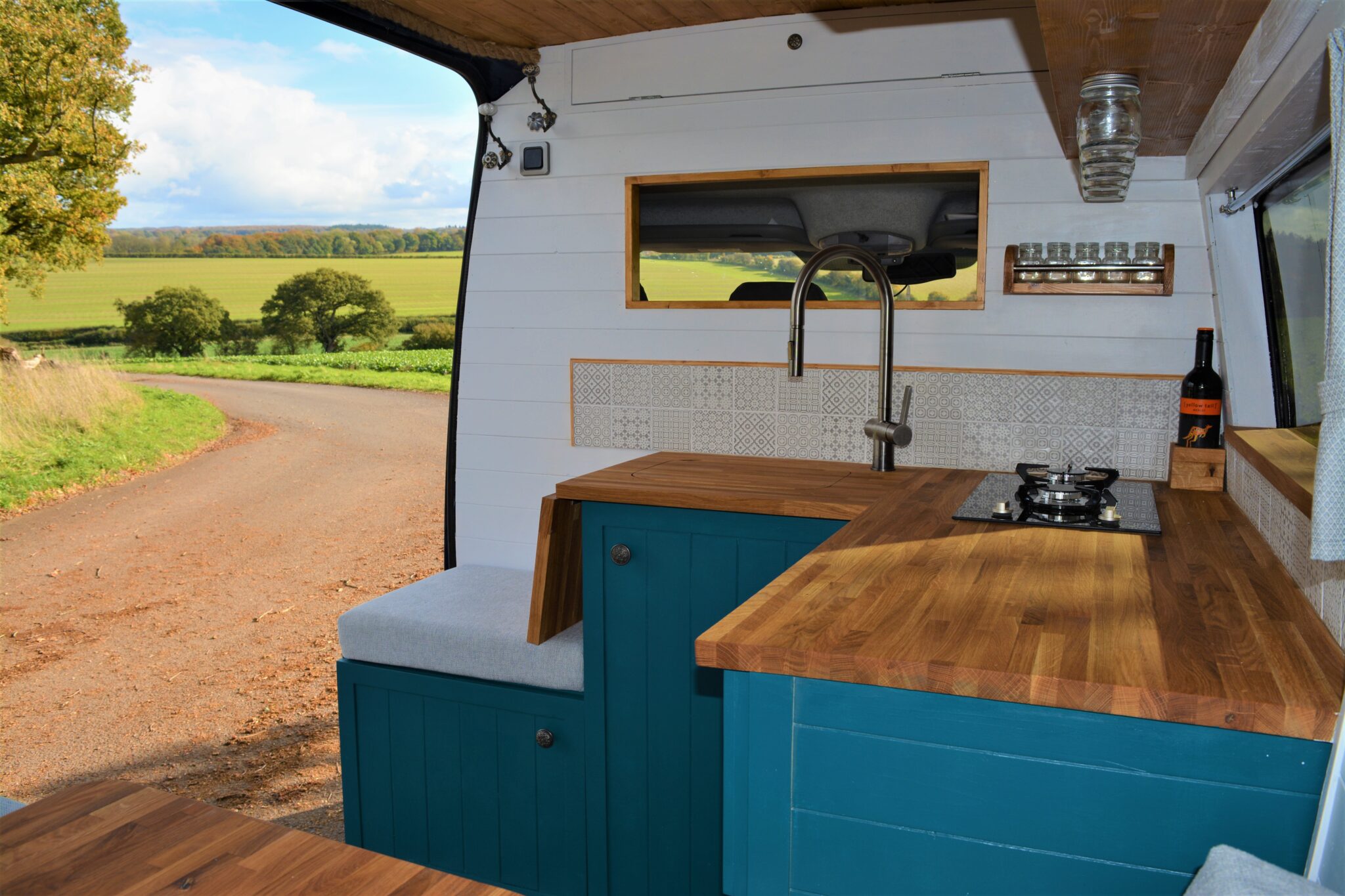 A cozy, rustic camper van interior featuring a wooden countertop, teal cabinetry, and a modern sink. Above the sink is a window with a picturesque view of a winding road and green fields. A small stove with two burners is set into the countertop, and a wine bottle is placed on the counter.