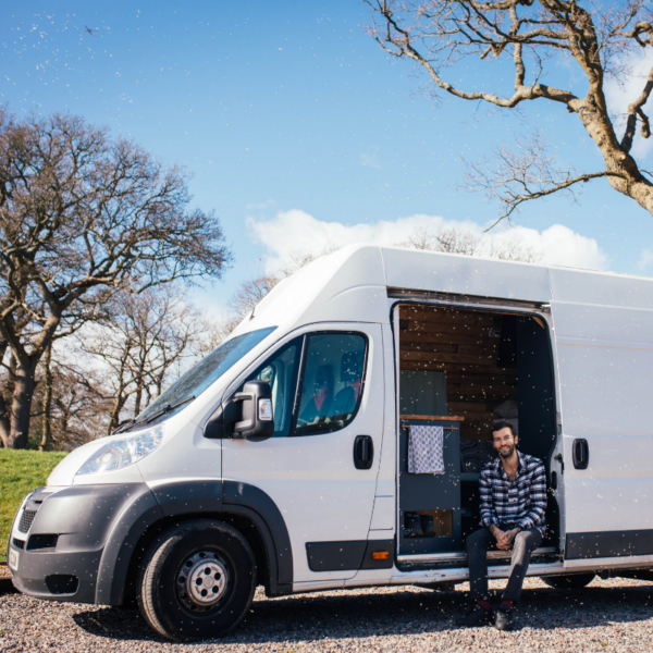 A white van with its side door open is parked on a gravel area near leafless trees. A man with a beard, wearing a black-and-white checkered shirt, sits on the van's step. The sky is blue with some clouds, and the surrounding area has green grass and bare branches, indicating early spring or late autumn.