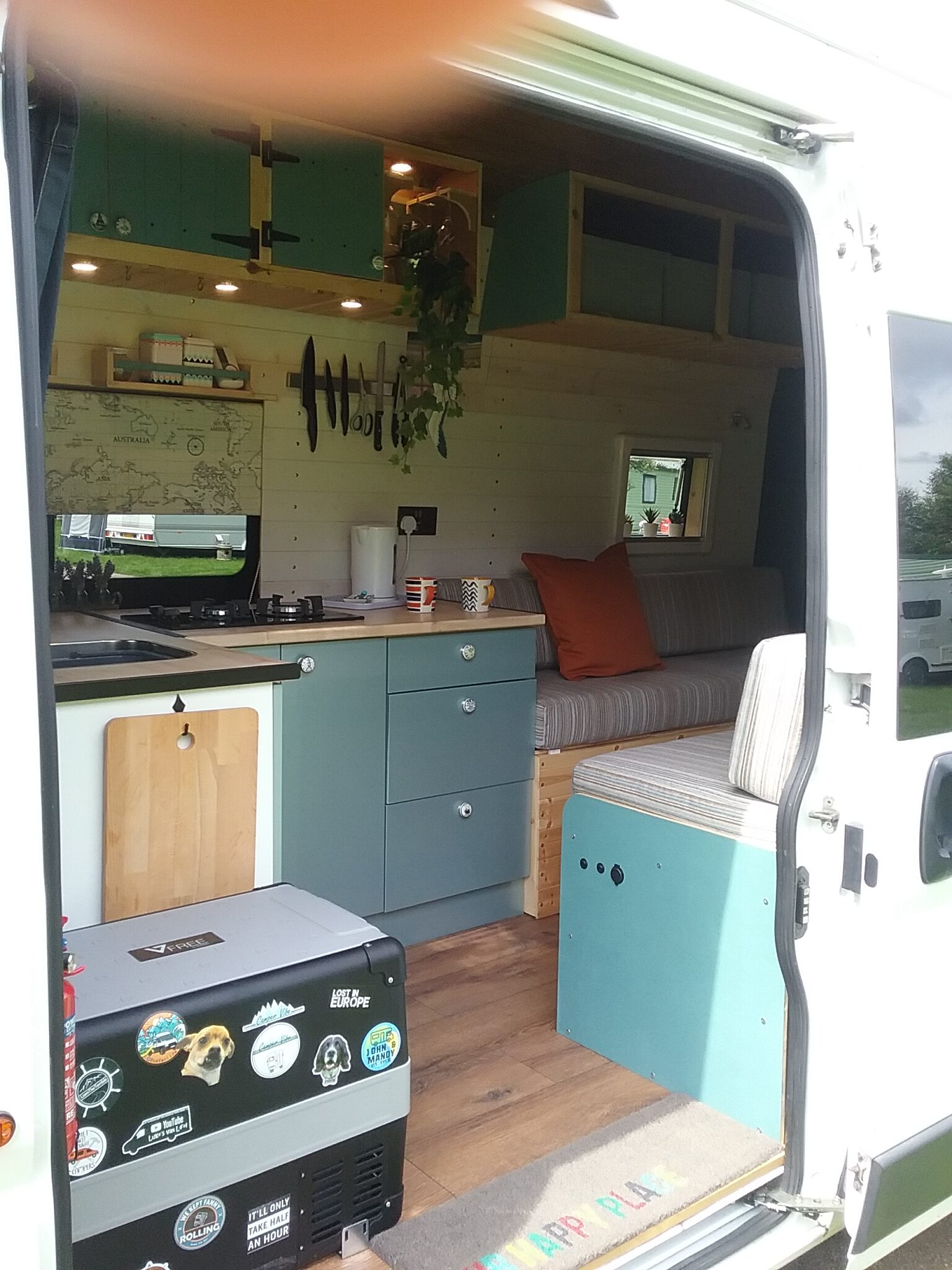 The interior of a camper van is shown with an open door. Inside, there's a small kitchenette with blue cabinets, a countertop with utensils hanging above, and a compact fridge adorned with stickers. A striped couch with orange and beige cushions sits beside a window. Some potted plants hang from the ceiling.