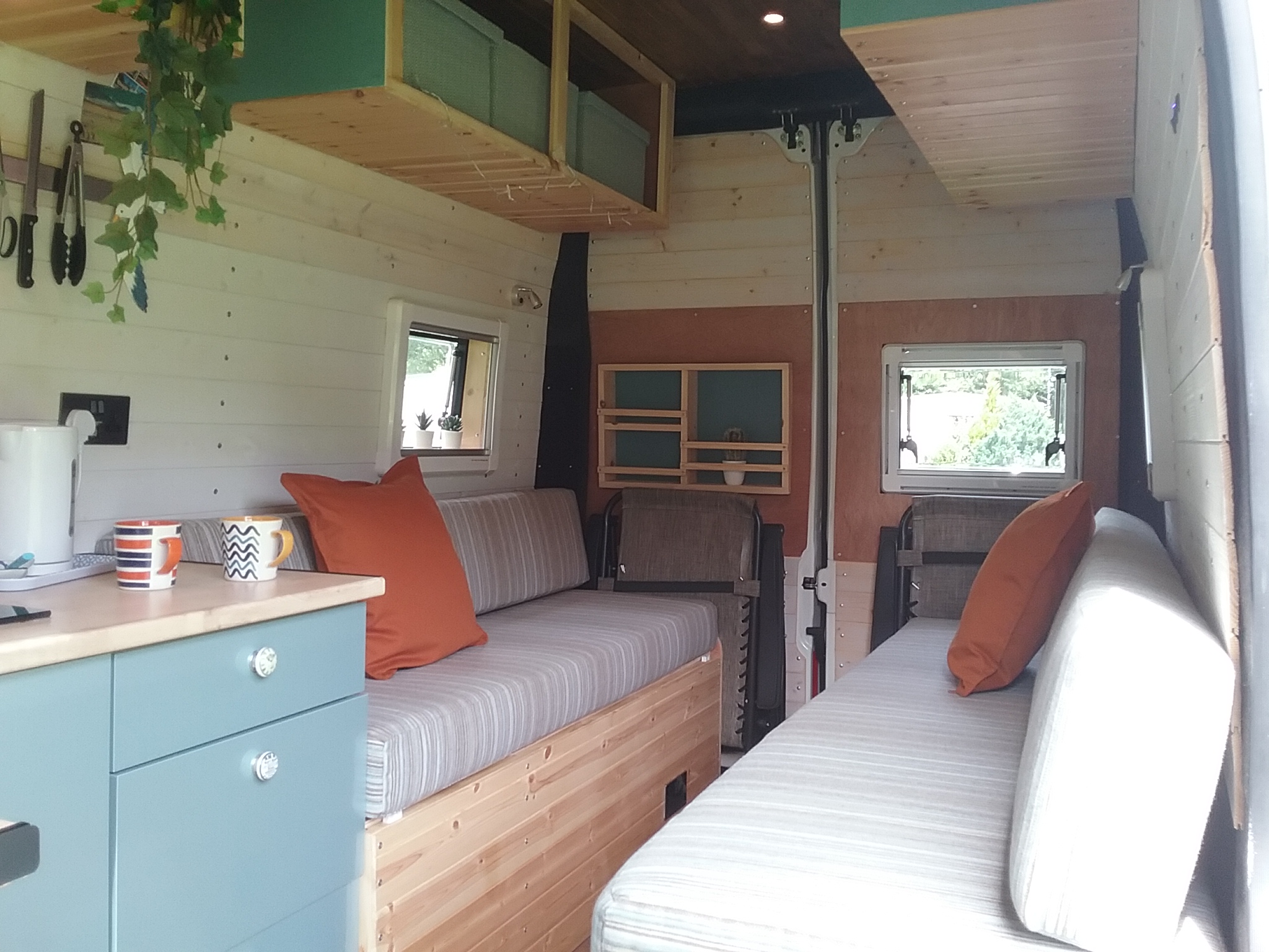 The interior of a converted camper van features two cushioned benches with orange pillows, a small kitchen area with cupboards and a countertop, hanging plants, and a window on each side. The walls and ceiling are finished in light wood. There is also a small set of shelves on the back wall.