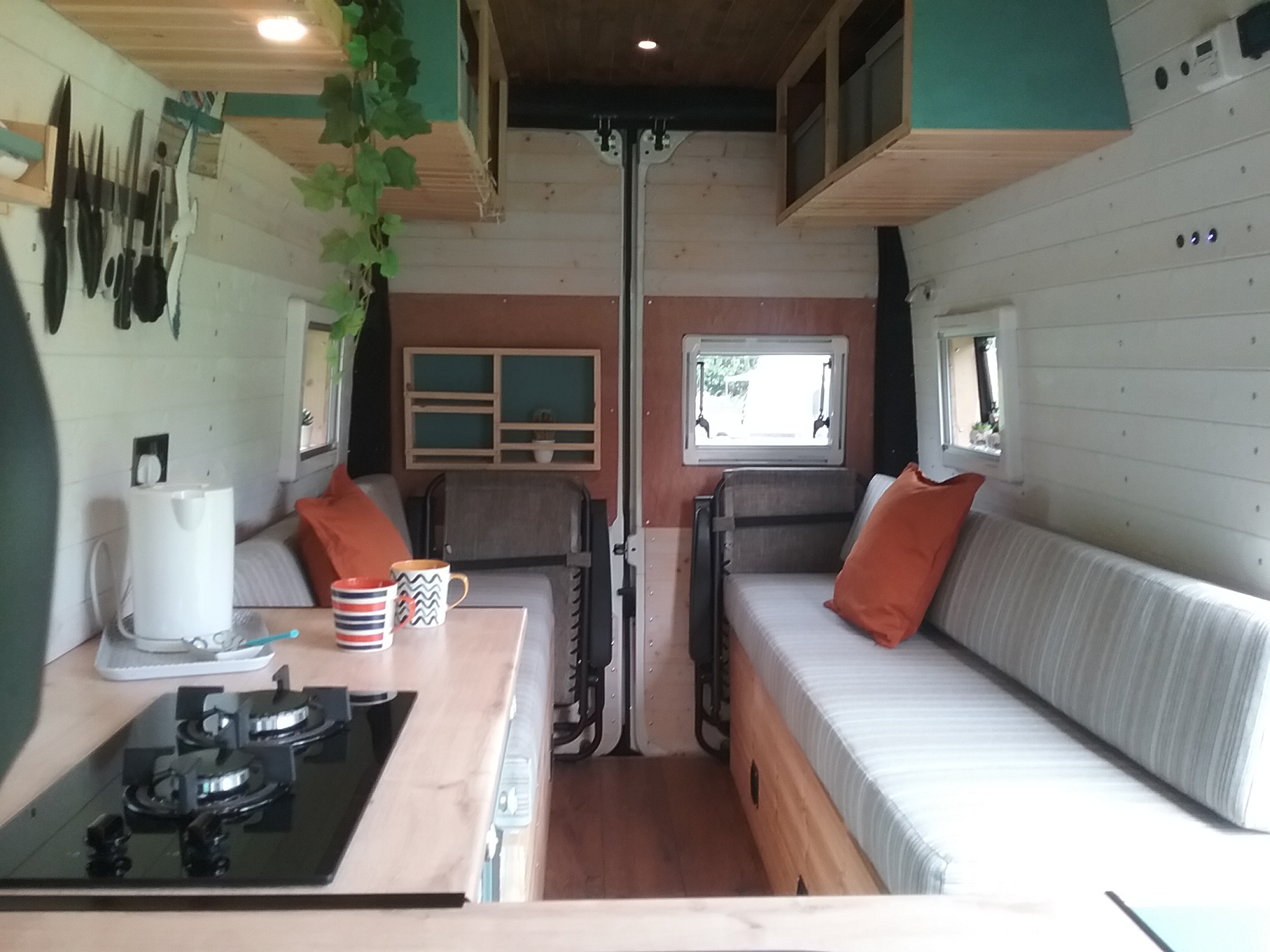Interior of a cozy van conversion featuring a kitchenette with a stove on the left, a striped sofa with orange cushions on the right, and small windows on both sides allowing natural light. The walls are lined with light-colored wooden panels, and there are hanging plants and shelves for storage.