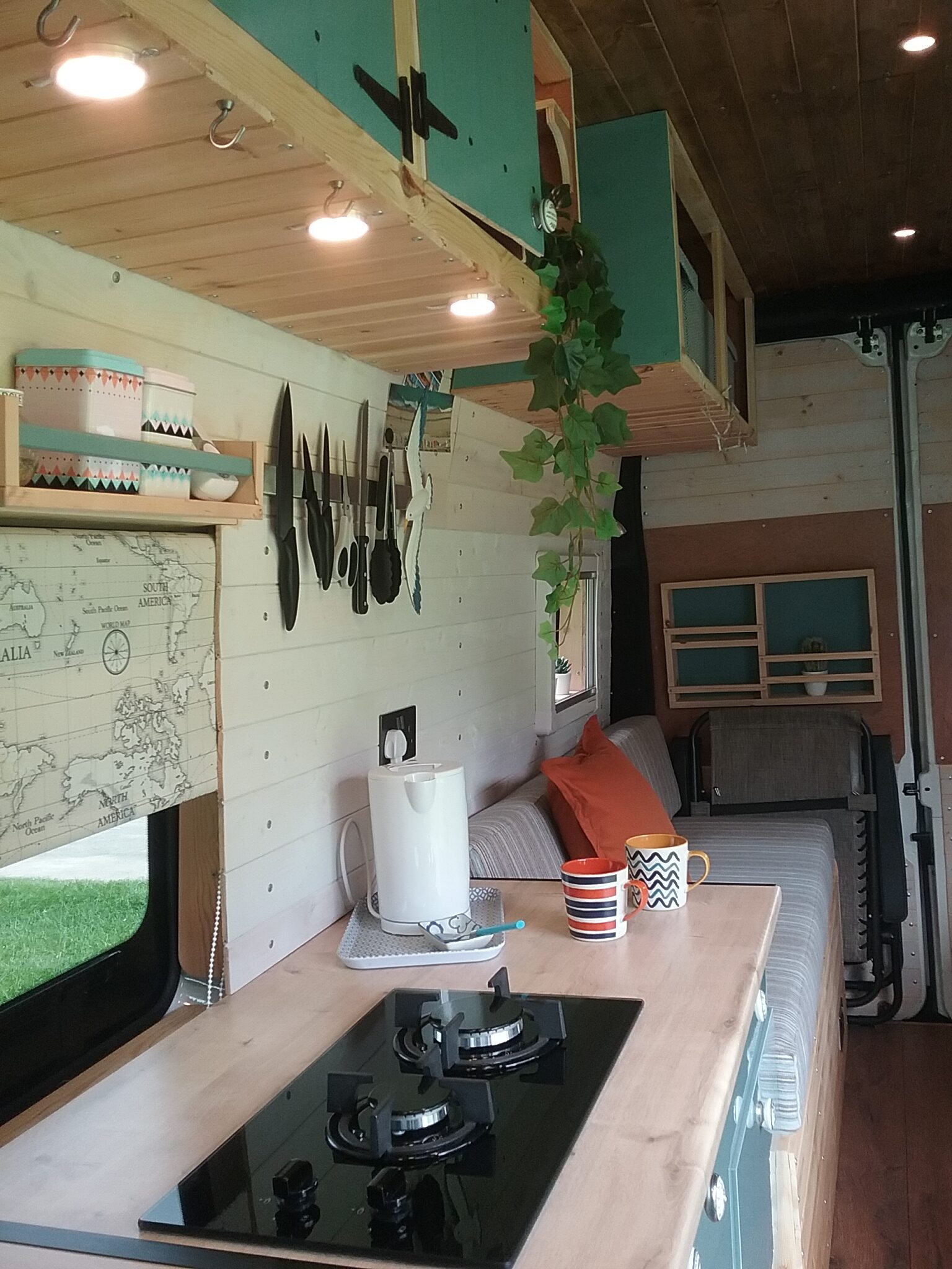 A cozy camper van interior with wooden paneling. It features a small kitchen area with a gas stove, kettle, and cutting board. Above, there are shelves with utensils and plants. The living space includes a cushioned bench with pillows and a map of Italy on the wall. Two mugs sit on the counter.