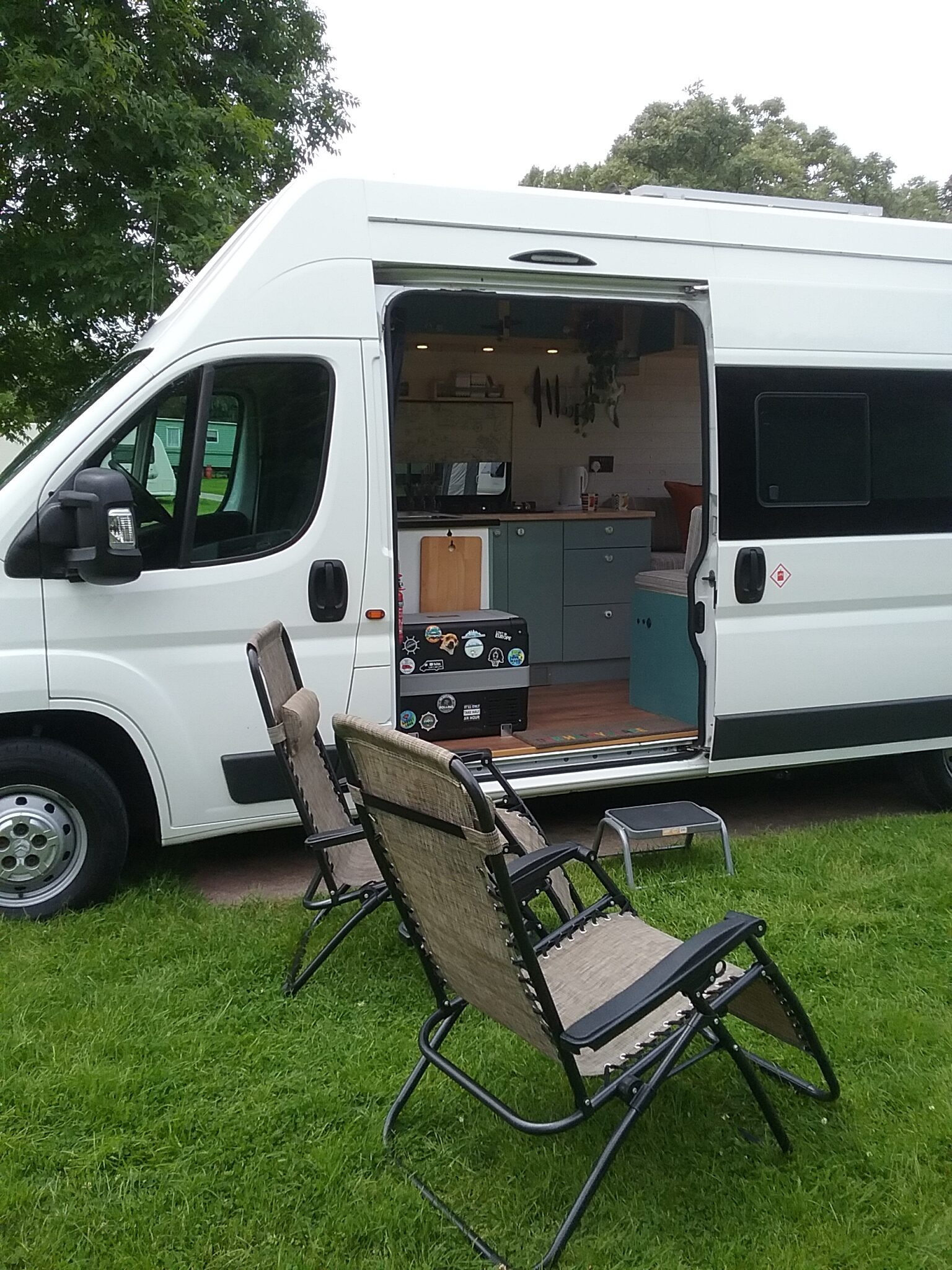 A white camper van with its side door open reveals a compact interior. Four folding chairs are set up on the grassy area outside. Inside the van are visible kitchen elements including a counter, sink, and cupboards. The scene depicts a cozy and organized mobile living space.