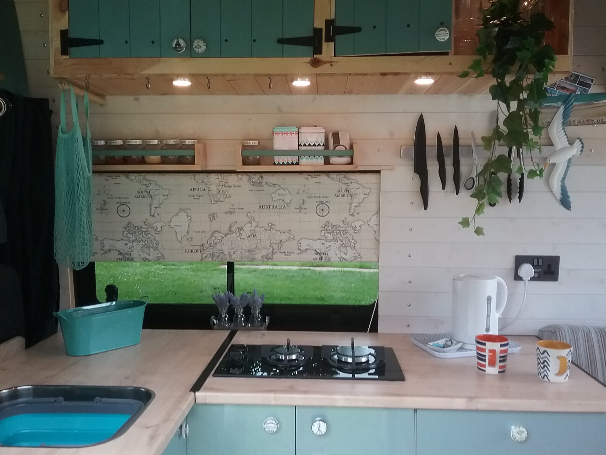 A cozy campervan kitchen features a two-burner stove, a sink, and a wooden countertop. Decor includes a world map window shade, small potted plants, teal and white storage cabinets, mugs, a kettle, and a hanging plant. Under-cabinet lights illuminate the tidy, well-organized space.