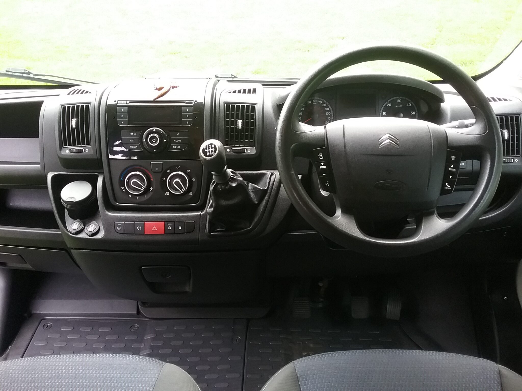 The image shows the interior of a Citroën vehicle's cockpit from the driver's perspective. It features a multifunction steering wheel, a central control panel with knobs and buttons for air conditioning and media, a gear stick, cup holders, and an instrument cluster behind the steering wheel.