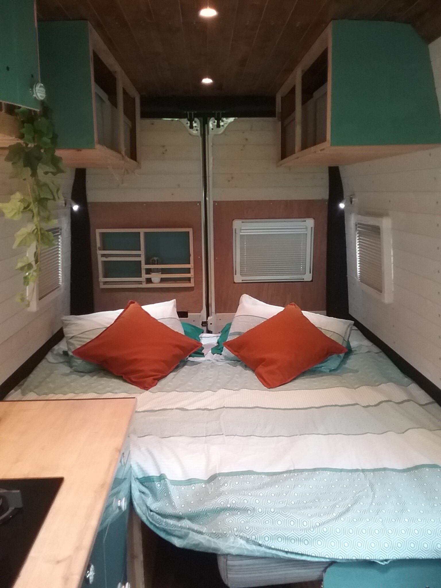 A cozy camper van interior featuring a neatly made bed with white and teal bedding. The bed is adorned with two white pillows and two orange cushions. Above the bed are teal overhead storage compartments. There is a small plant hanging from the upper left corner, and a wooden countertop is partially visible on the left side.