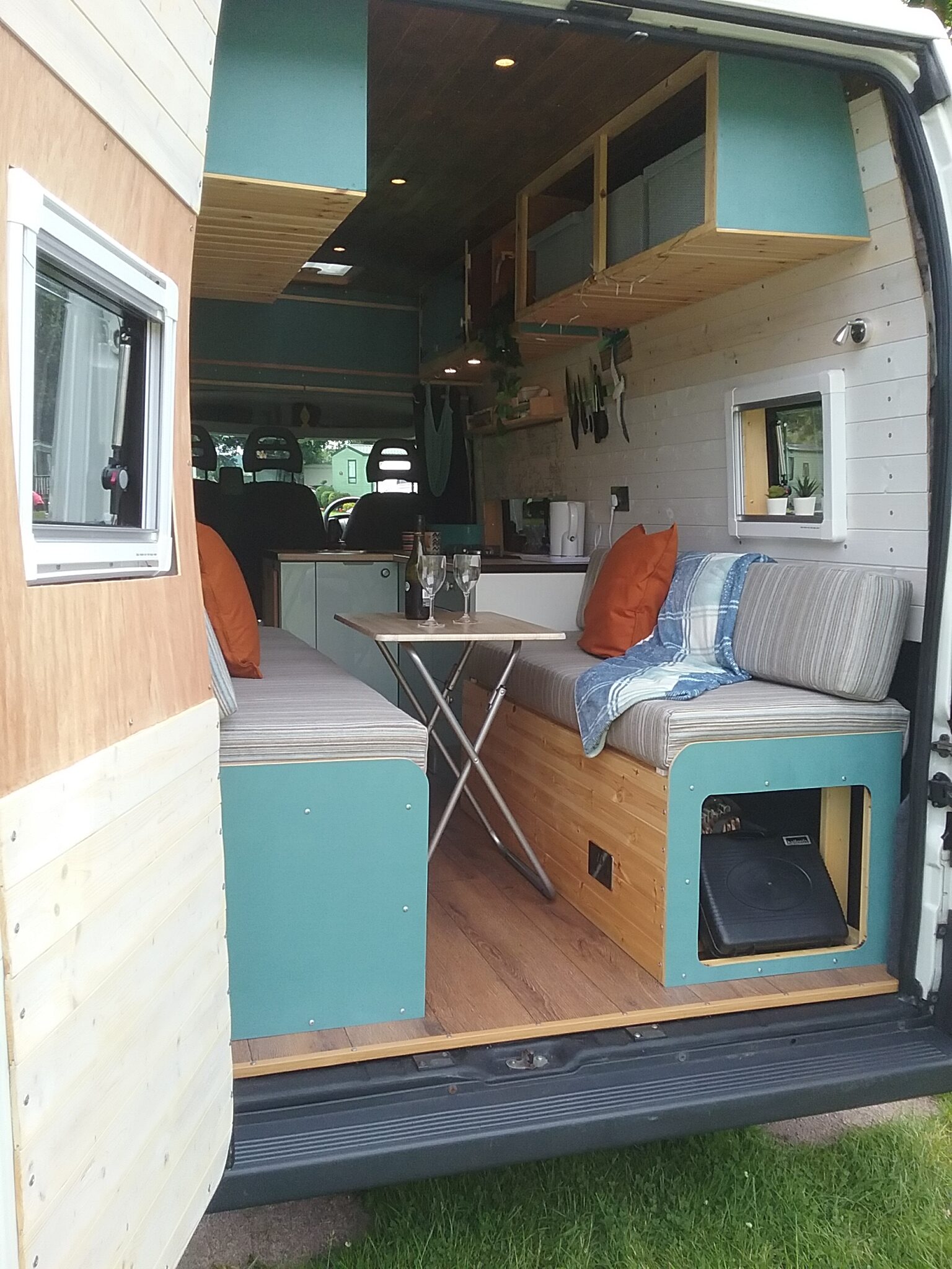 A converted camper van with the rear doors open reveals a cozy interior. The space includes two cushioned bench seats with orange throw pillows, a folding table in between, and overhead storage cabinets. The wooden paneling and plants add a homey touch. A front cab area is visible beyond the living space.