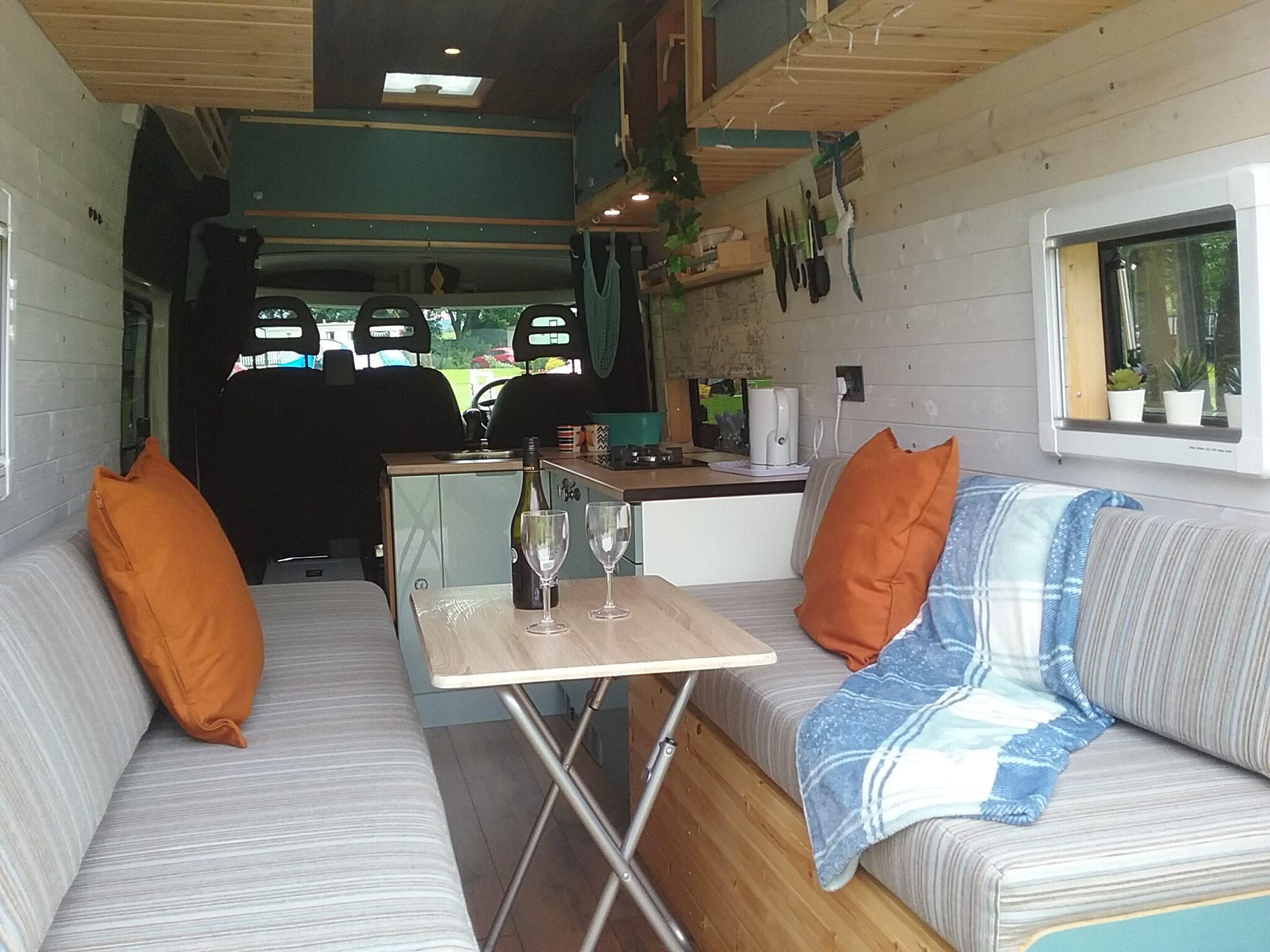 Interior of a cozy converted camper van featuring a seating area with striped cushions, orange pillows, and a blue-checked blanket. A foldable table holds two glasses and a wine bottle. The kitchen has hanging utensils and storage cabinets. A window with potted plants offers natural light.