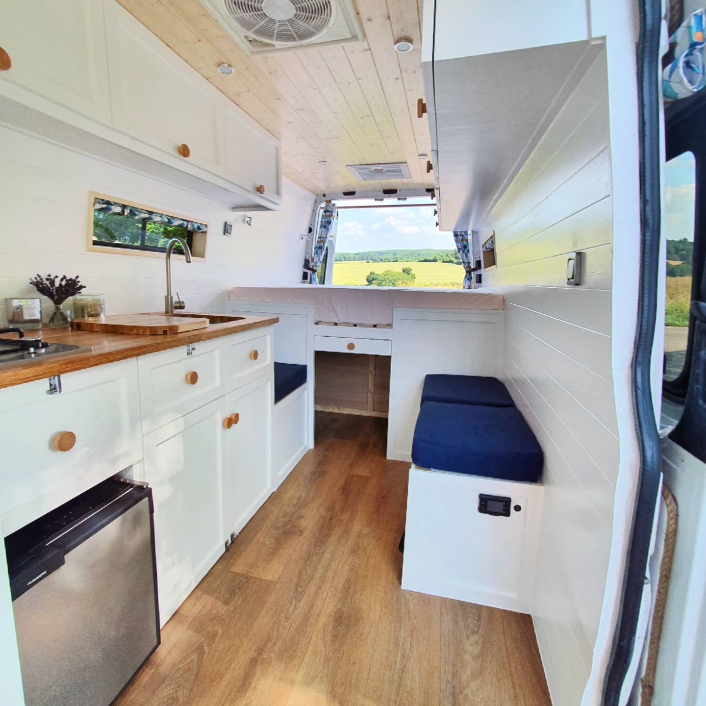 Interior of a converted van with white wooden cabinets, a small stainless steel sink, a stovetop, and a wood countertop. Dark blue cushions on benches flank a fold-out table. White wood paneling lines the walls and ceiling. A rear door frames a pastoral landscape of fields and distant trees.