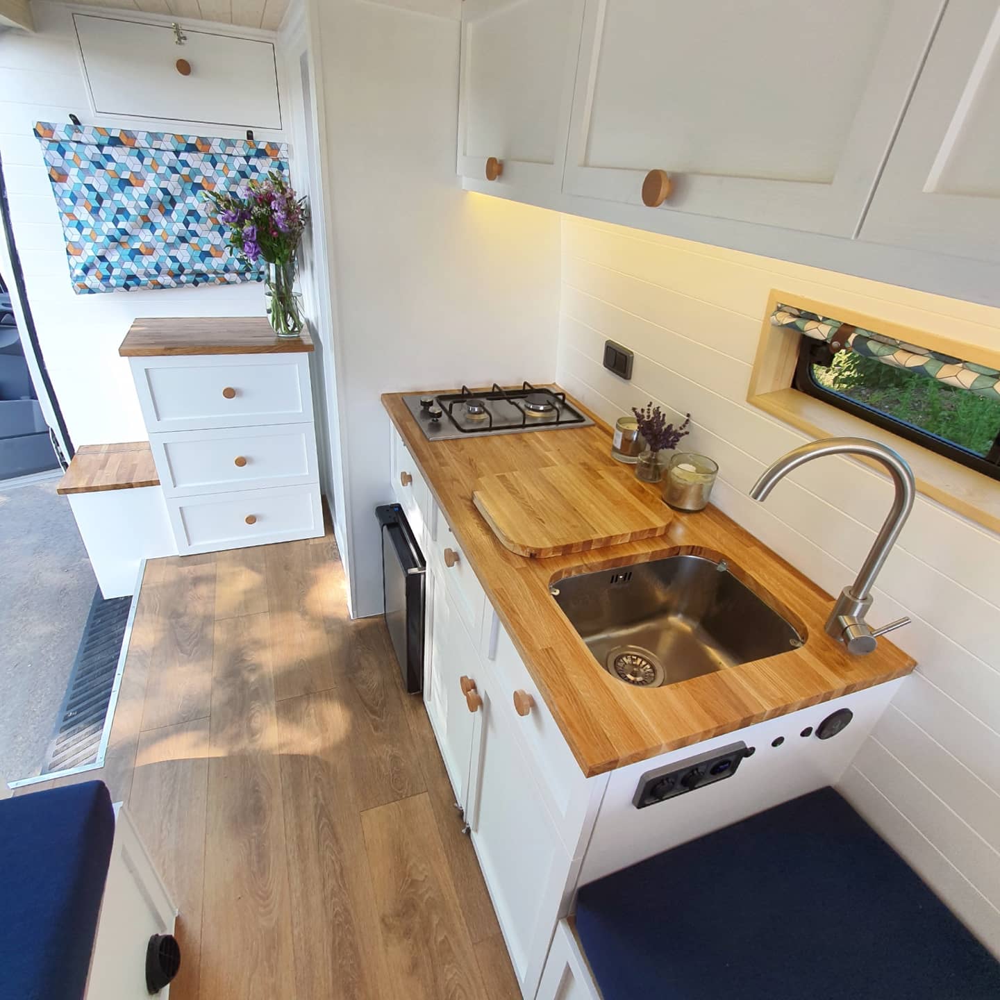 A compact kitchen area in a mobile home features a wooden countertop with a built-in stainless steel sink and a two-burner gas stove. White cabinetry, including drawers and cupboards, provides storage. A small potted plant and a cut board are on the counter, and there’s a bench with drawers nearby.