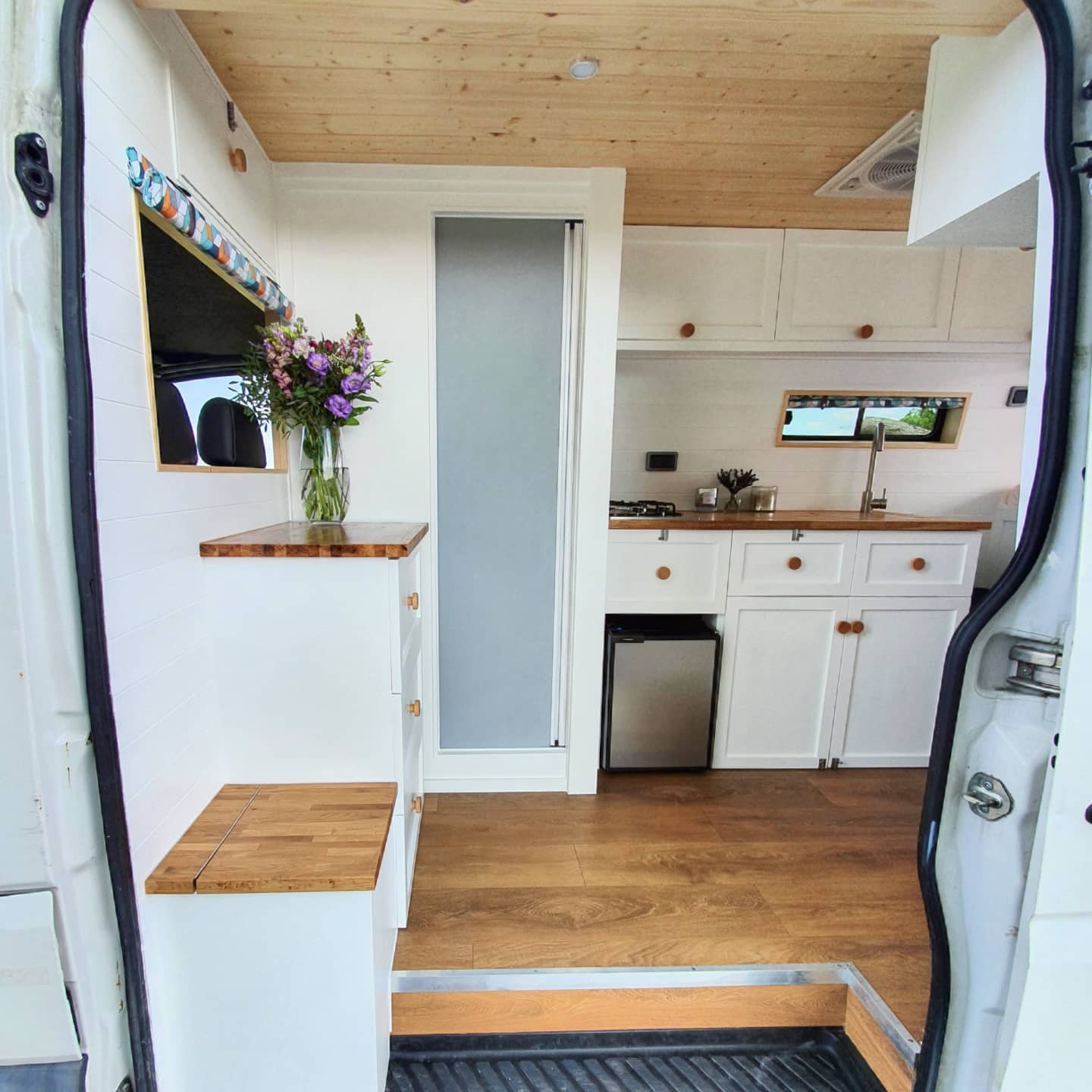 Interior of a cozy, converted camper van. White cabinetry and wooden countertops create a clean look. A small fridge sits below the counter. Flowers in a glass vase decorate a ledge on the left. A stove is built into the counter, and a window above the sink brings in natural light.