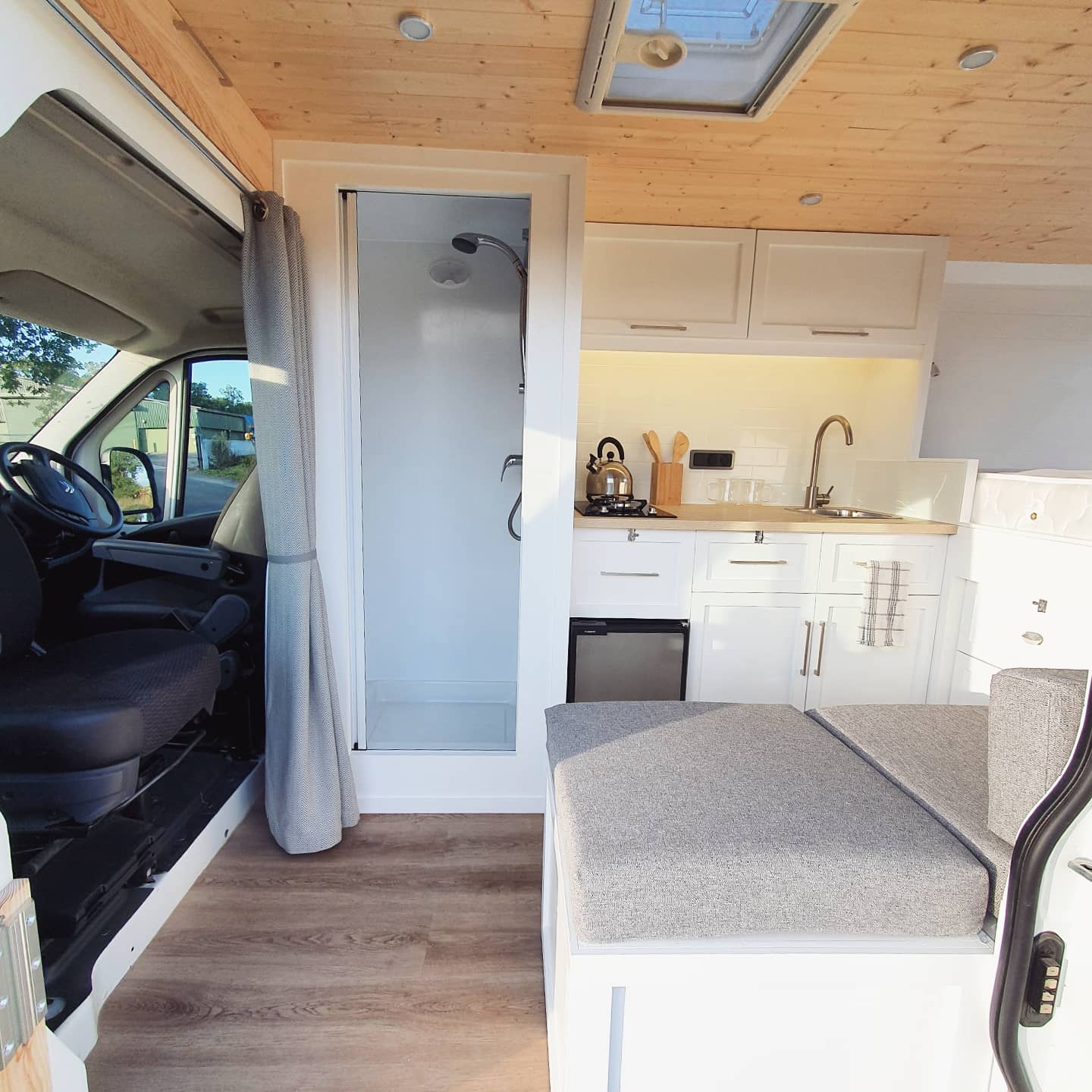 Interior view of a camper van featuring a compact kitchen and shower. The kitchen includes a small fridge, sink, counter space with a kettle, cutting boards, and light-colored cabinetry. Adjacent is a shower stall with a curtain. The foreground has a cushion seat and visible wooden flooring.