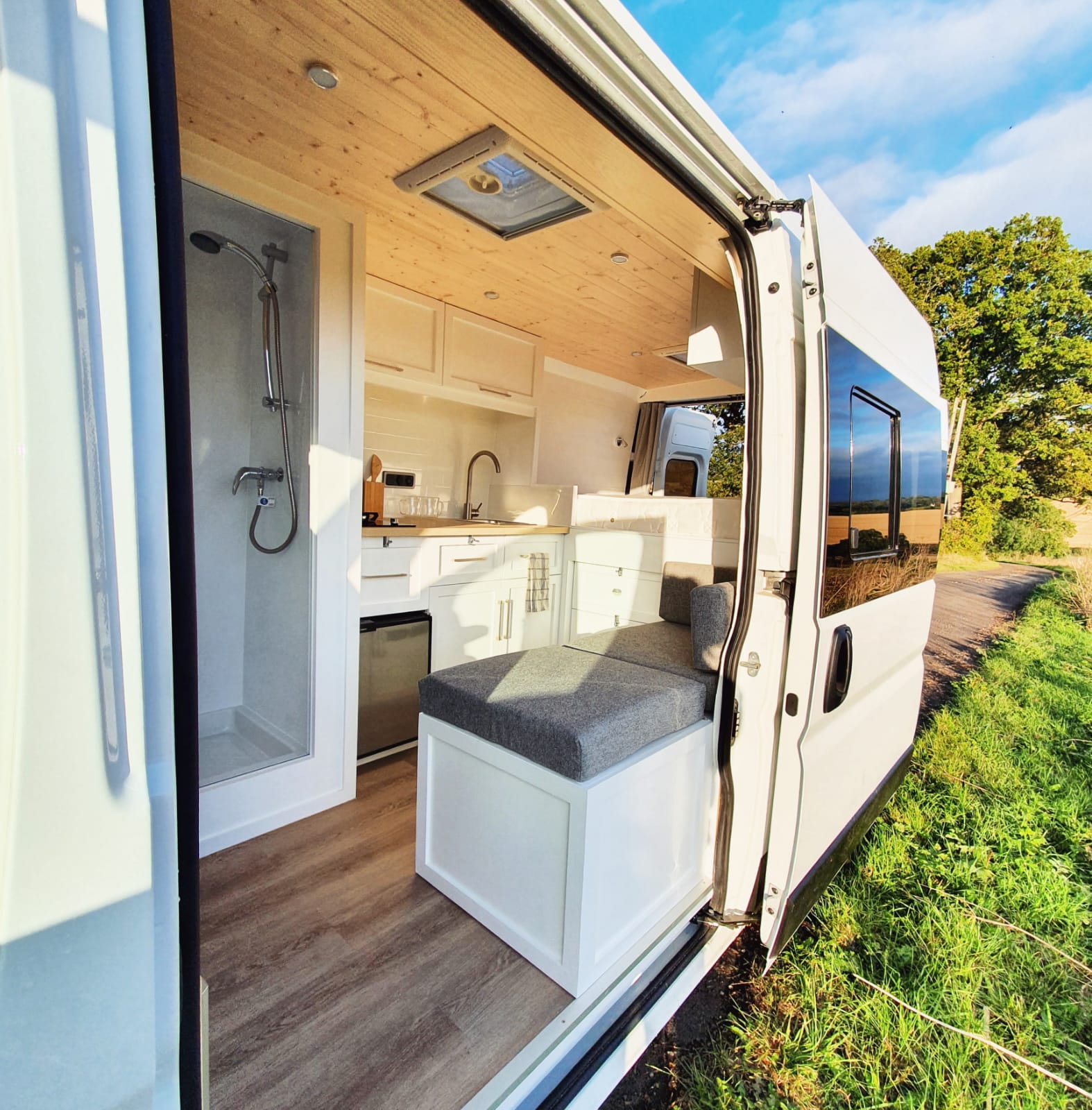 A bright and modern camper van with its side door open, revealing a compact yet cozy interior. Inside, there is a small kitchenette with a sink, stove, and refrigerator. Adjacent to the kitchen is a cushioned bench seat. The ceiling and upper cabinets are made of light-colored wood.