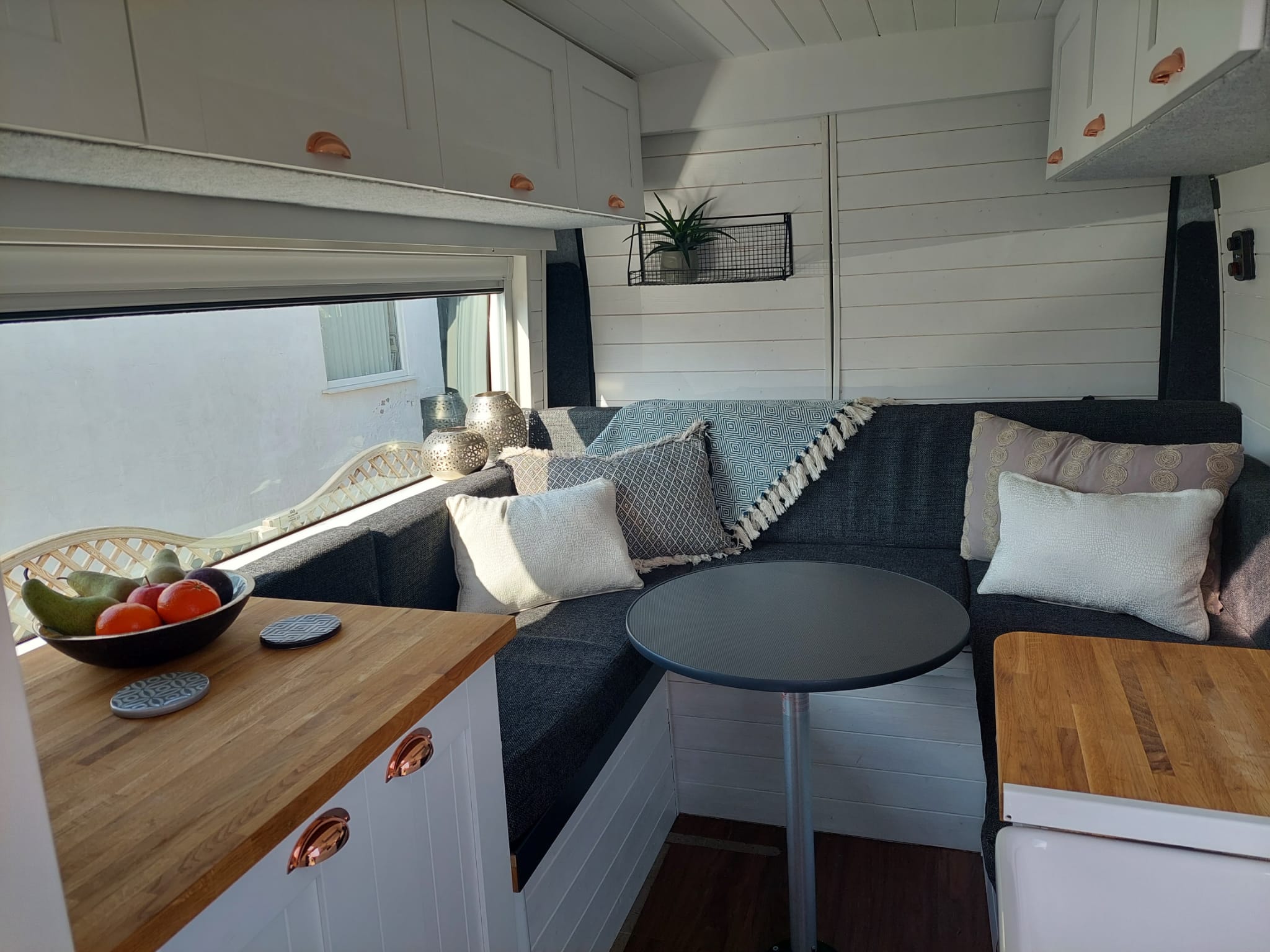 A cozy RV interior with a U-shaped seating area featuring dark cushions and various throw pillows. A small, round black table is centered in front of the seating. Adjacent to the seating is a wooden counter. A large window lets in natural light, illuminating the space.