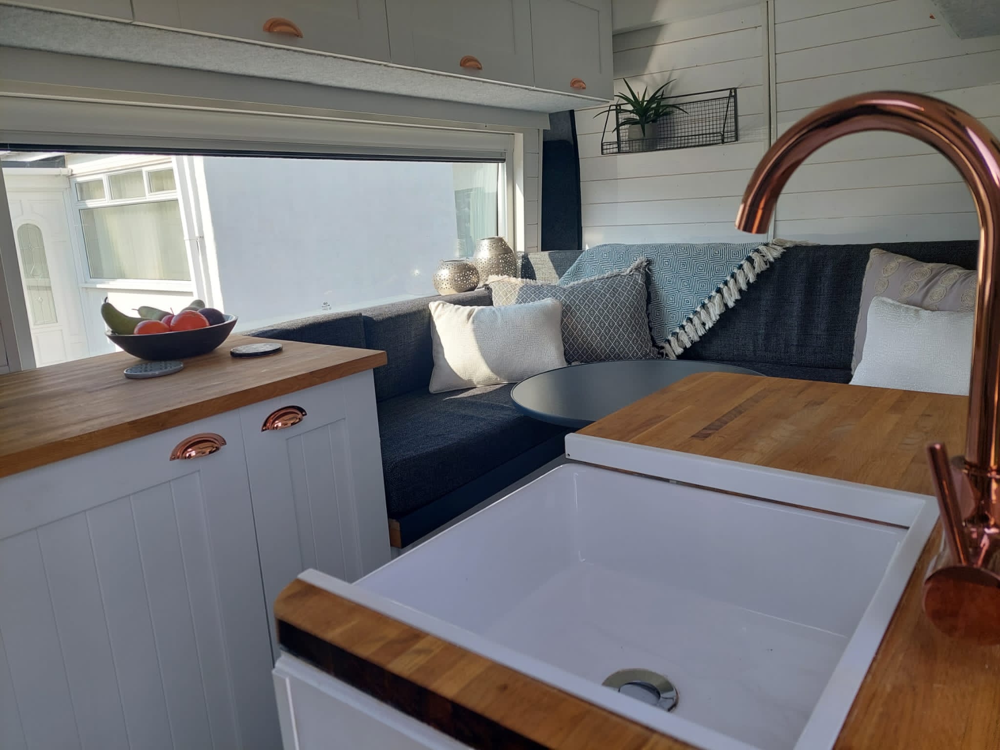A cozy RV interior featuring a white kitchen with a wooden countertop and a copper faucet. A white sink is in the forefront. The seating area includes gray and blue cushions, a small round table, and a wooden bench. A large window brings in natural light, and a plant decorates the back wall.