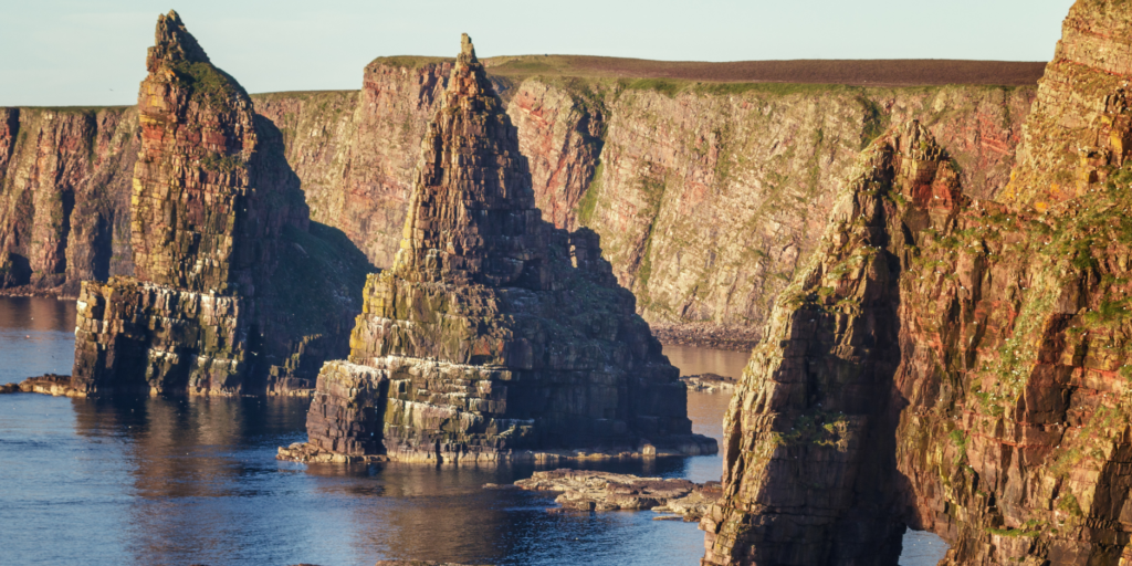 Steep, rocky sea stacks rise dramatically from the calm ocean waters along a rugged coastline with multi-coloured cliffs. The formations are jagged and weathered, surrounded by deep blue water, with lush, grassy cliff tops stretching into the distance under a clear sky.