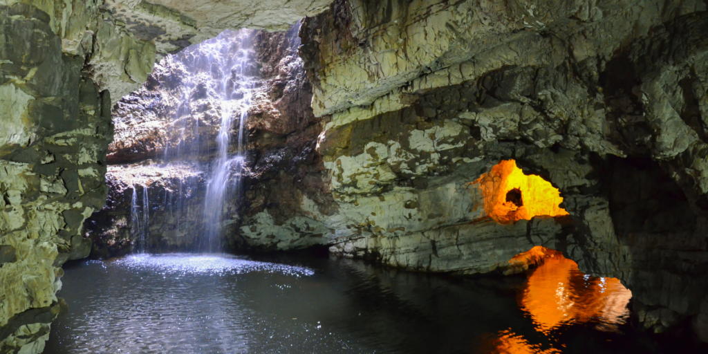 A picturesque cave scene with a waterfall cascading from an opening in the rocky ceiling into a serene pool of water. There’s an illuminated archway on the right-hand side, casting a warm, golden glow that reflects off the water, contrasting with the cool, natural light of the falling water.