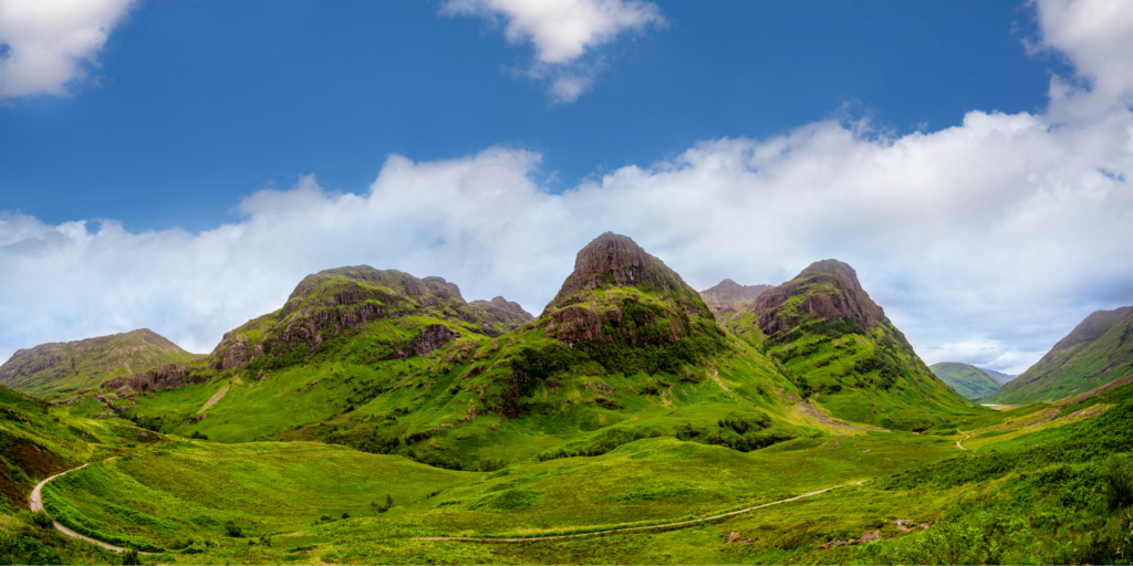 Panoramic view of the scenic Three Sisters of Glencoe in the Scottish Highlands, featuring three contiguous hills covered in lush green grass and dotted with rocky outcrops, under a partly cloudy blue sky. A meandering path winds through the verdant valley in the foreground.
