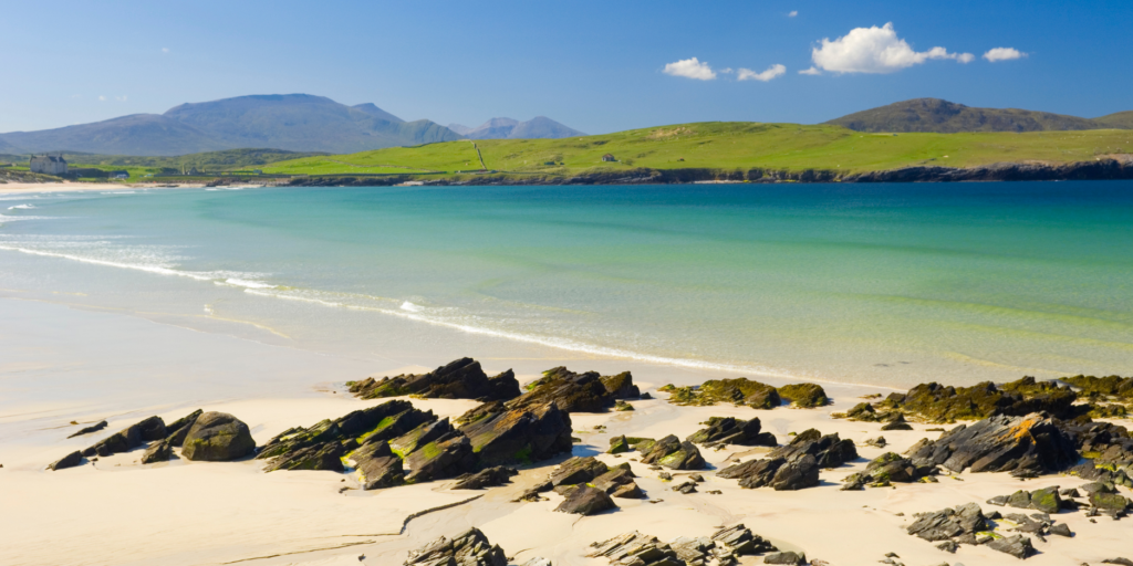 A picturesque beach scene showcases golden sand and jagged rocks in the foreground. Turquoise waters gently lap against the shore, extending out to meet lush green hills and mountains under a bright blue sky with a few scattered clouds.