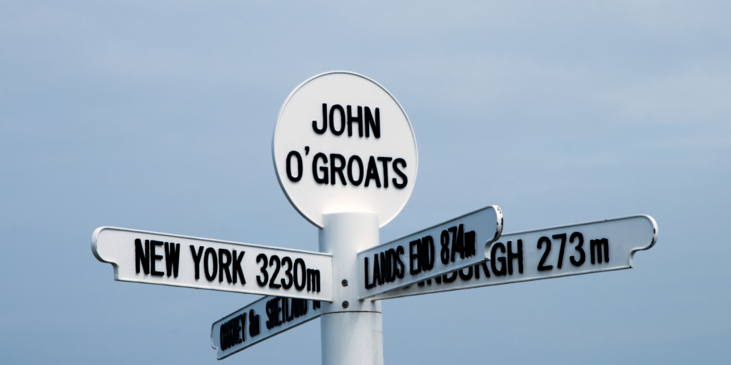 A white signpost at John O'Groats stands against a cloudy sky, adorned with numerous directional arms. One arm points to New York, 3230 miles away; another directs towards Land's End at a distance of 874 miles; and yet another to Edinburgh, which is 273 miles away. At the top of the sign, "John O'Groats" is boldly displayed in black letters