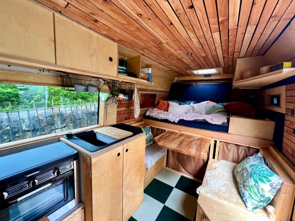 Bonnie offers a cosy camper van interior with wood panelling,. It features a compact kitchen with a hob and sink on the left, a small bed adorned with colourful cushions at the back, open shelving, and bench seating with storage underneath. A window provides a serene view of greenery outside, enhancing the overall ambience.